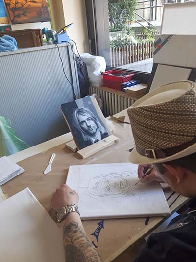 Der Künstler sitzt am Tisch und zeichnet das Portrait von Curt Cobain. Er ist von hinten zu sehen mit einen Dandy-Strohhut auf dem Kopf.