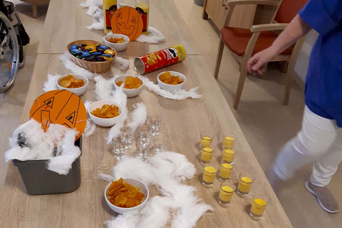 Tisch mit Halloween-Dekoration wie einem Papierkürbis mit gruseligem Gesicht, Gläser mit Eierlikör, Süßes und Chips.