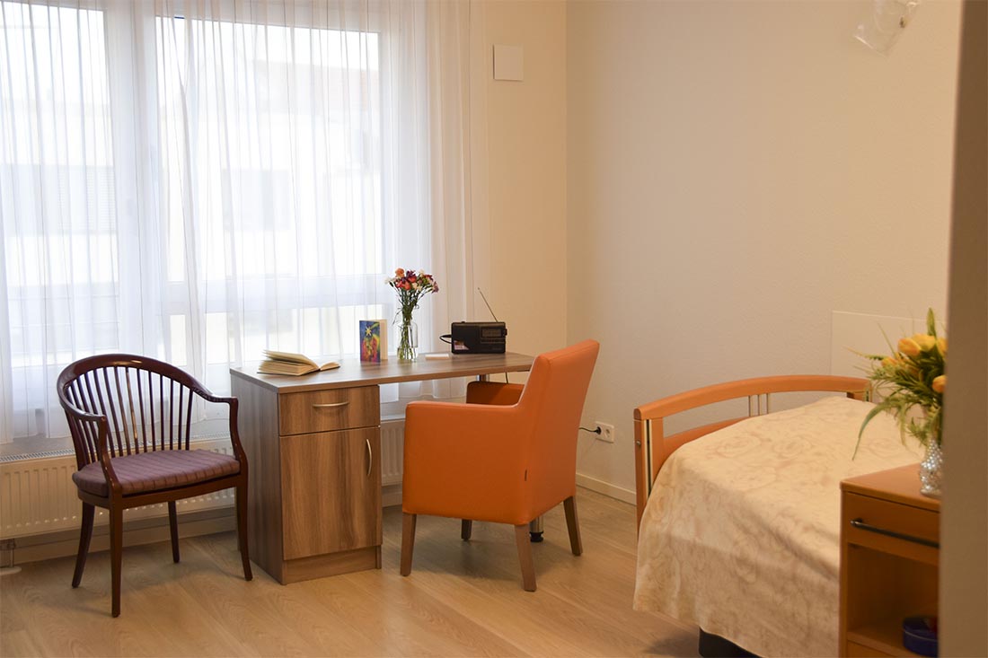 Modernes Zimmer im Pflegeheim, mit Schreibtisch, Stuhl und einem orangenen Sessel vor dem Schreibtisch. Rechts im Bild ist ein Stück des Pflegebetts zu sehen.